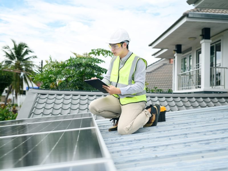 A young Asian male technician in safety vest and hard hat kneels on a rooftop inspecting solar panels with a clipboard, ideal for renewable energy and sustainability concepts.