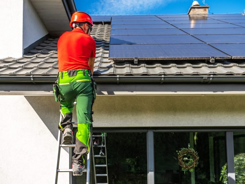 A worker wearing a hard hat and safety gear is climbing a ladder to install solar panels on a residential roof. The panels are already partially installed and cover a significant portion of the roof.