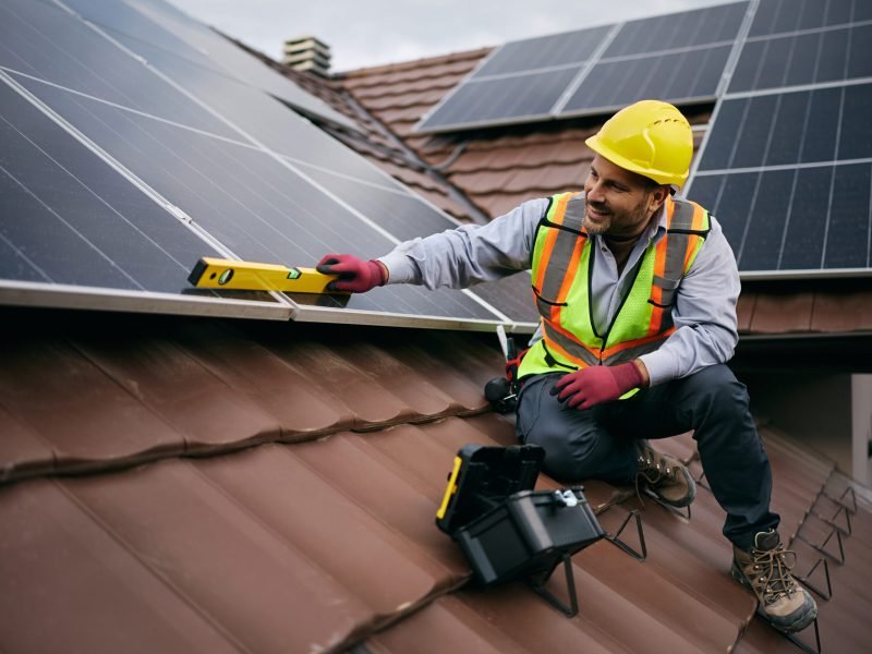 Happy worker using level while installing solar panels on the rooftop. Copy space.